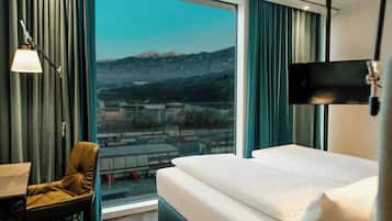 A hotel room with a bed, a desk, a chair, a TV, and a large window with a view of a city and mountains.
