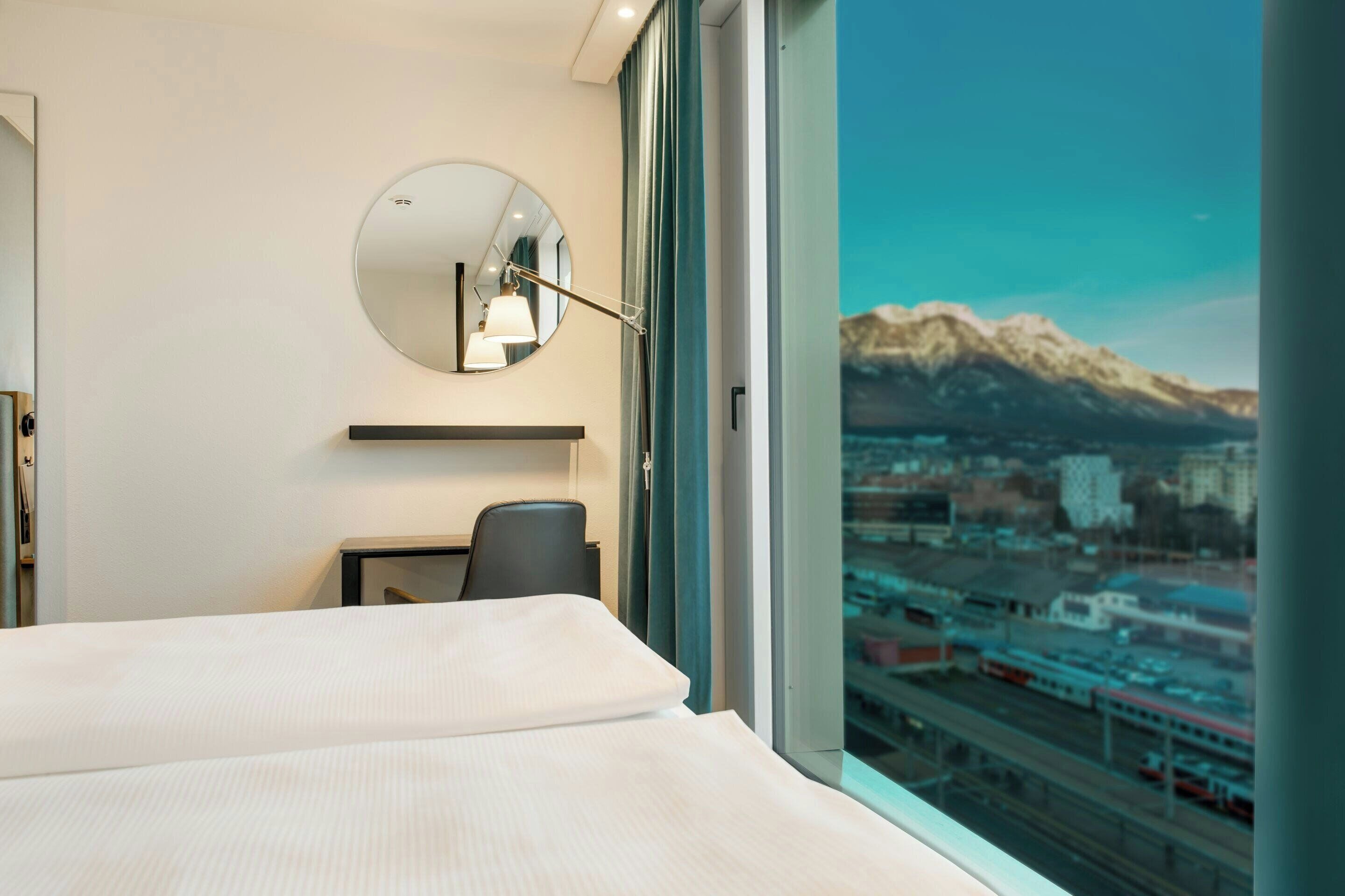 Une chambre d’hôtel comprenant un lit, un bureau, une chaise, un miroir rond et une grande fenêtre donnant sur la montagne.