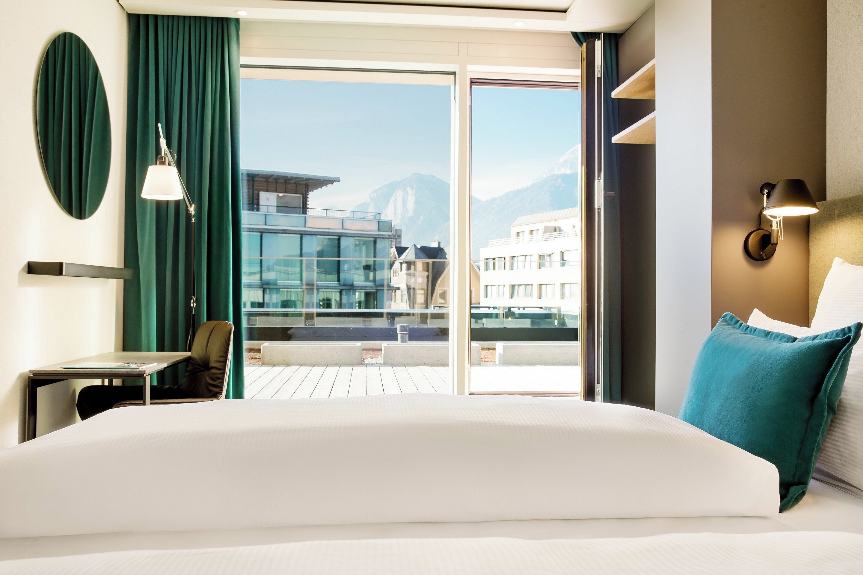 Quarto de hotel com uma cama grande, uma escrivaninha, uma cadeira, uma janela com vista para edifícios e montanhas, e uma varanda.