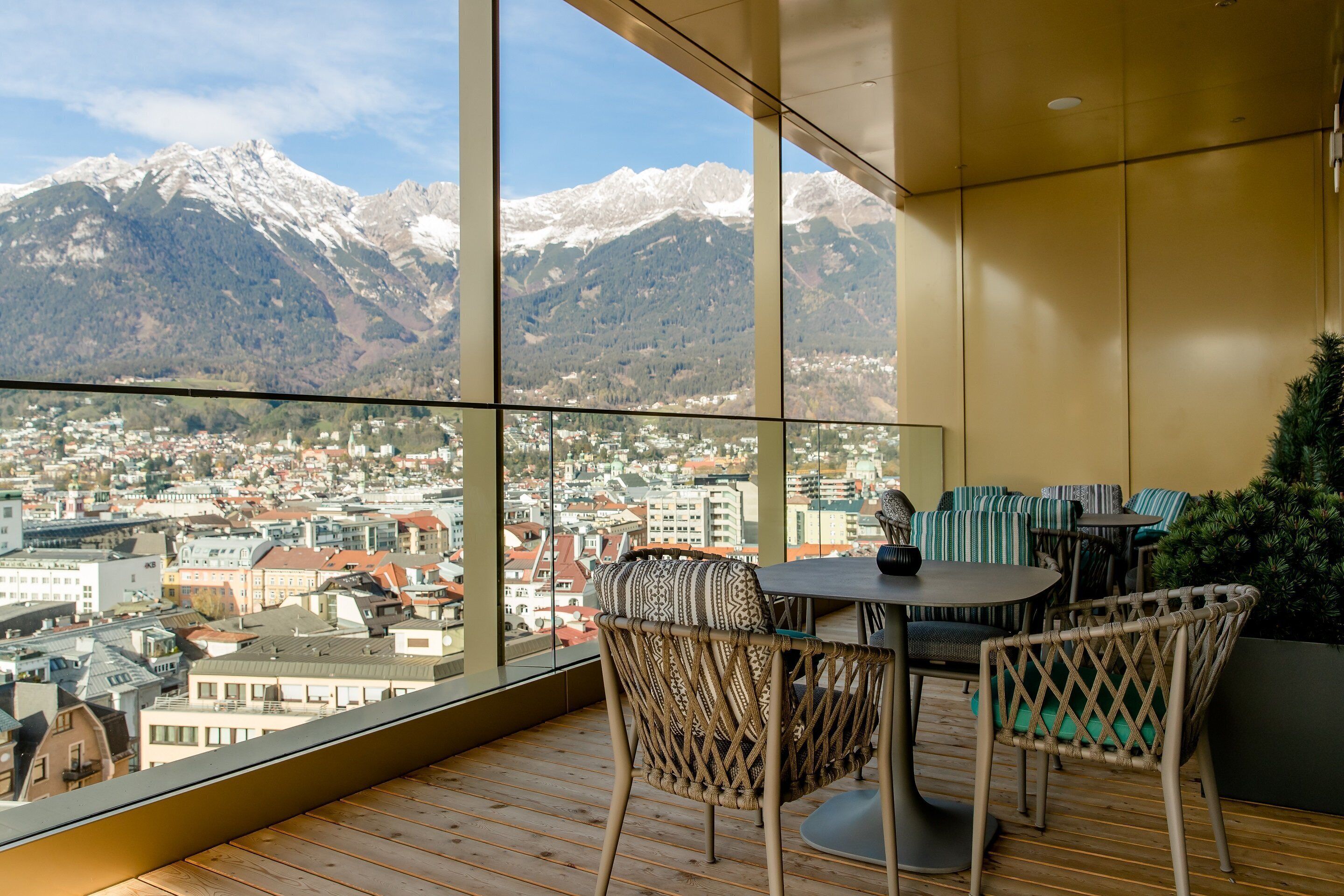 Ein Balkon mit Blick auf eine Stadt und Berge. Korbstühle und ein Tisch mit einer Vase sind vorhanden.