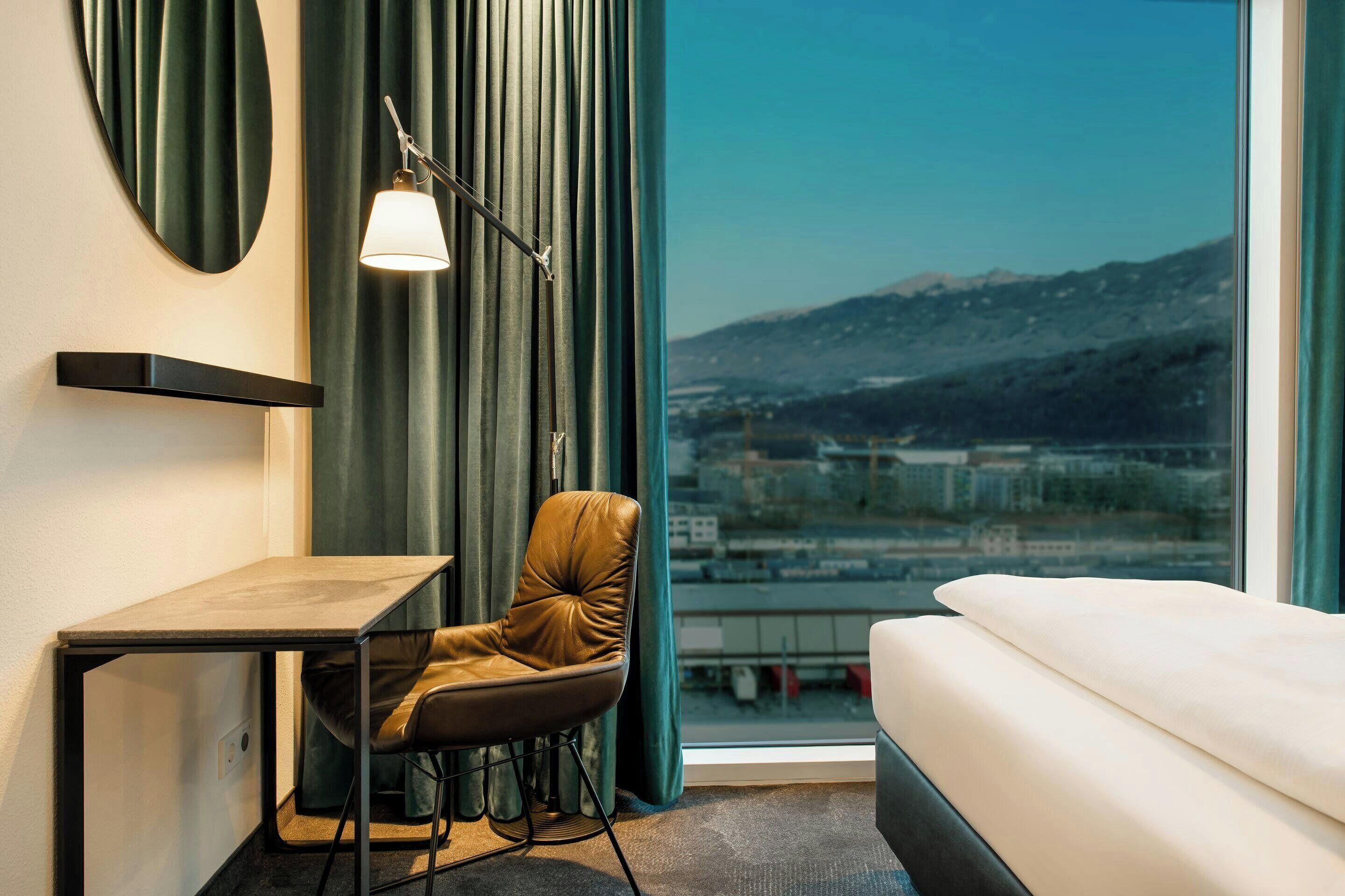 Habitación de hotel con cama, escritorio, silla, lámpara y vista panorámica de la ciudad a través de una ventana.