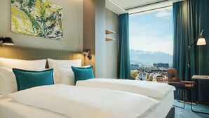 A hotel room with two beds, a desk, a chair, a lamp, and a large window with a city and mountains view. - Motel One Innsbruck (Innsbruck)