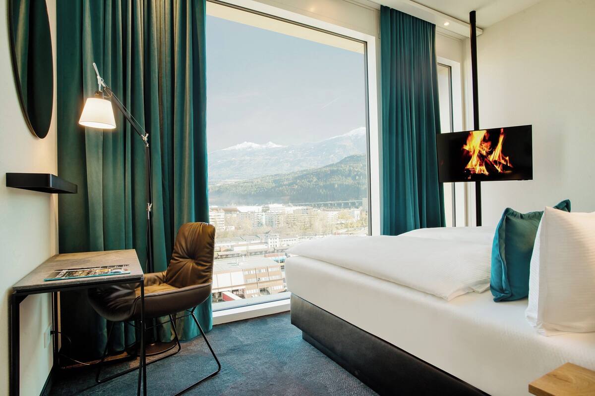 A hotel room with a bed, a desk, a chair, a TV, and a view of a cityscape.