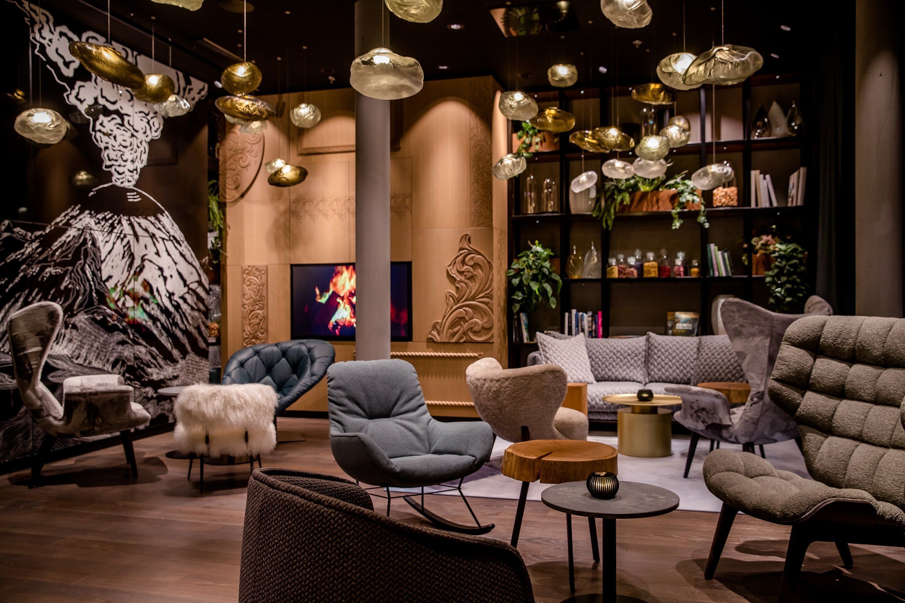 Un interior moderno con diversas opciones de asiento, incluyendo un sillón gris, un otoman blanco de pelo suave y una mecedora negra. Se observa una estantería llena de libros y objetos decorativos, así como un televisor montado en la pared.