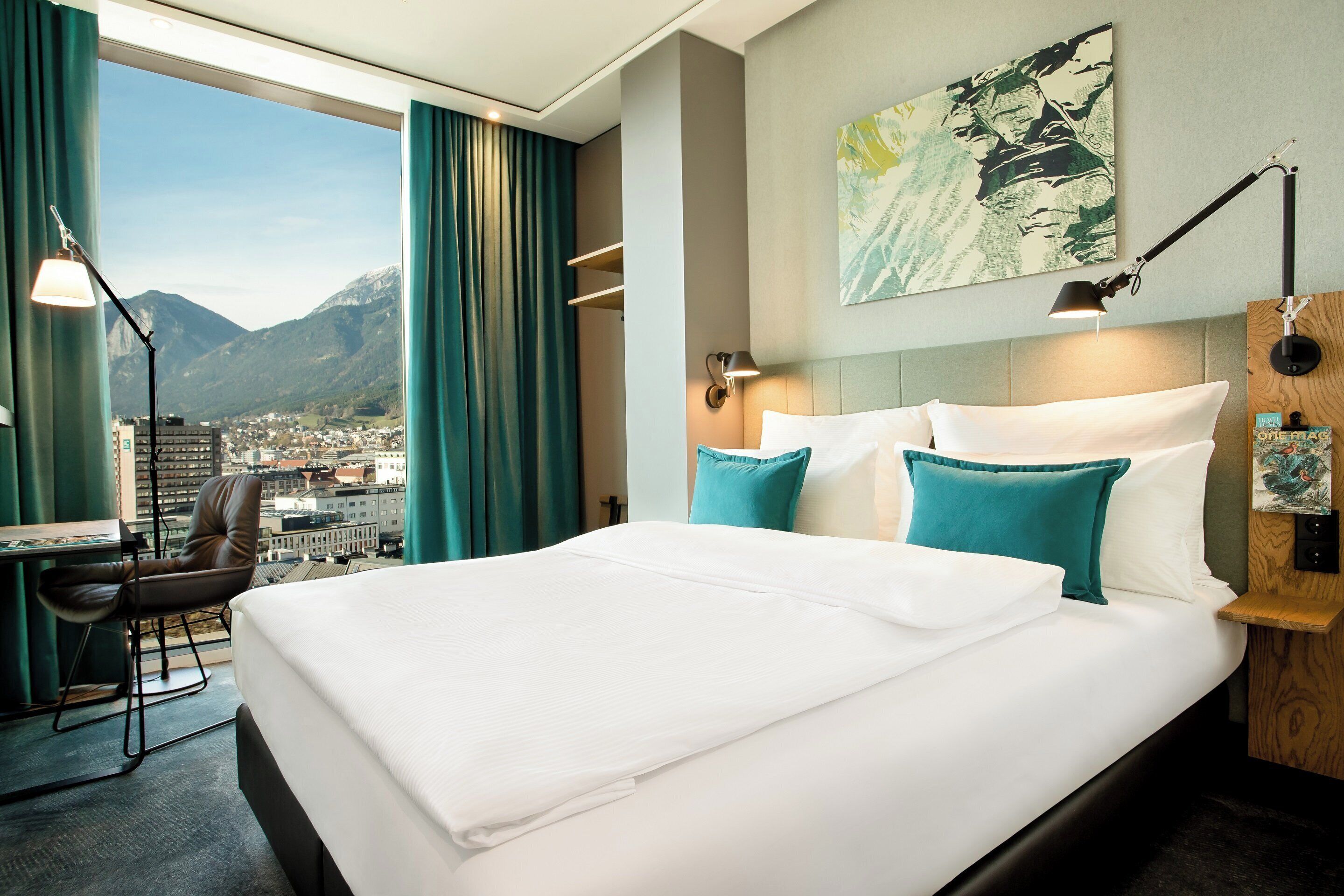 A hotel room with a large bed, a desk, and a chair. There is a view of mountains and a cityscape through the window.