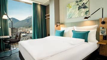 Une chambre d’hôtel avec un grand lit, un bureau et une chaise. La fenêtre offre une vue sur les montagnes et sur le paysage urbain.