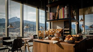 A cozy café with a wooden counter, a variety of breads and pastries, hanging pendant lights, and a bookshelf filled with books. - Motel One Innsbruck (Innsbruck)