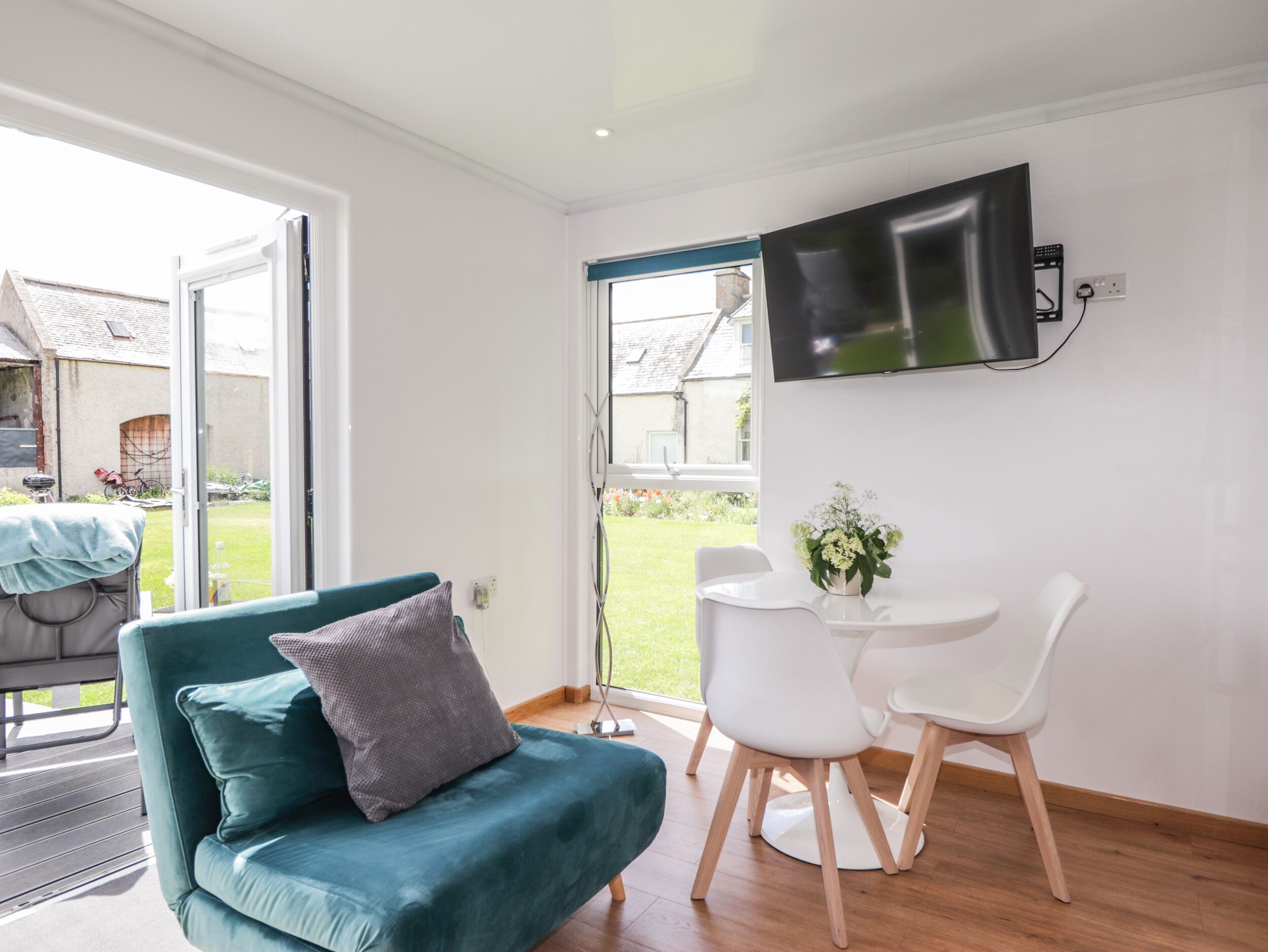Ein modernes Wohnzimmer mit einem türkisblauen Sofa, einem weißen Esstisch mit zwei Stühlen, einem an der Wand befestigten Flachbildfernseher und einem großen Fenster mit Blick auf den Garten.