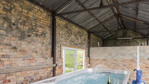 A hot tub with water jets, a bottle of champagne, and two glasses, set against a brick wall with a metal roof. - Moss of Bourach (Aberlour)