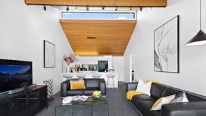 Un salon moderne avec un téléviseur à écran plat, une table basse en verre et un canapé en cuir noir. Il y a également un coin cuisine avec des meubles blancs et un plafond en bois.