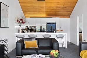 A modern living room with a black leather sofa, a coffee table with magazines, and a fruit bowl. A kitchen with white cabinets and a large window is visible in the background. - STUNNING LAKE VIEW LUXURY ALPINE VILLA (Queenstown)