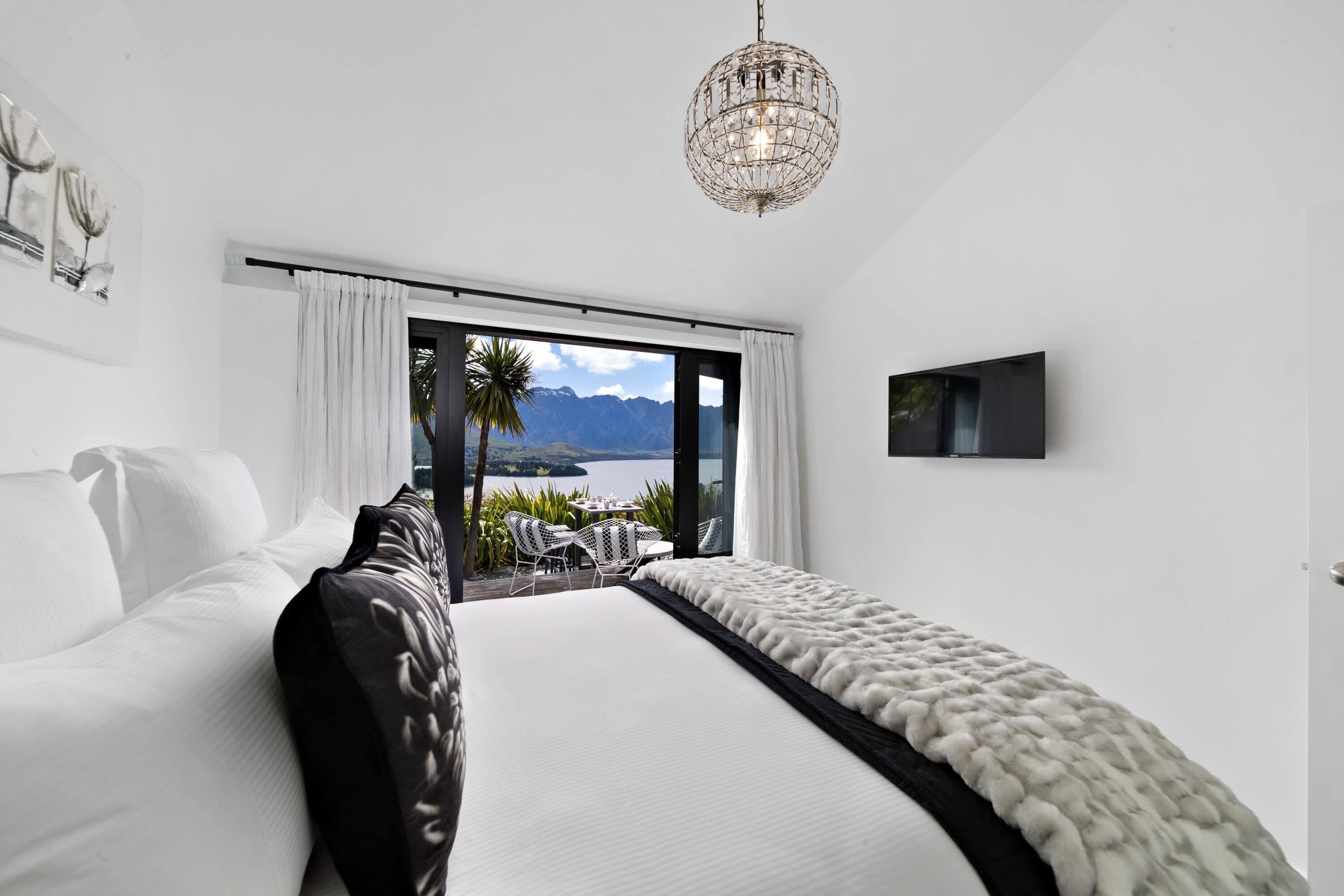 A modern bedroom with a large bed, a view of mountains and water, a chandelier, and a flat-screen TV.