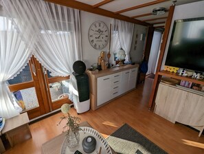 Ein gemütliches Wohnzimmer mit Holzboden, einer großen Wanduhr, einem Flachbildfernseher und einer Essecke mit Tisch und Stühlen.