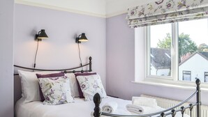 Una cama bien tendida con sábanas blancas y almohadas florales, dos lámparas de lectura fijadas a la pared y una ventana con vistas a una casa.