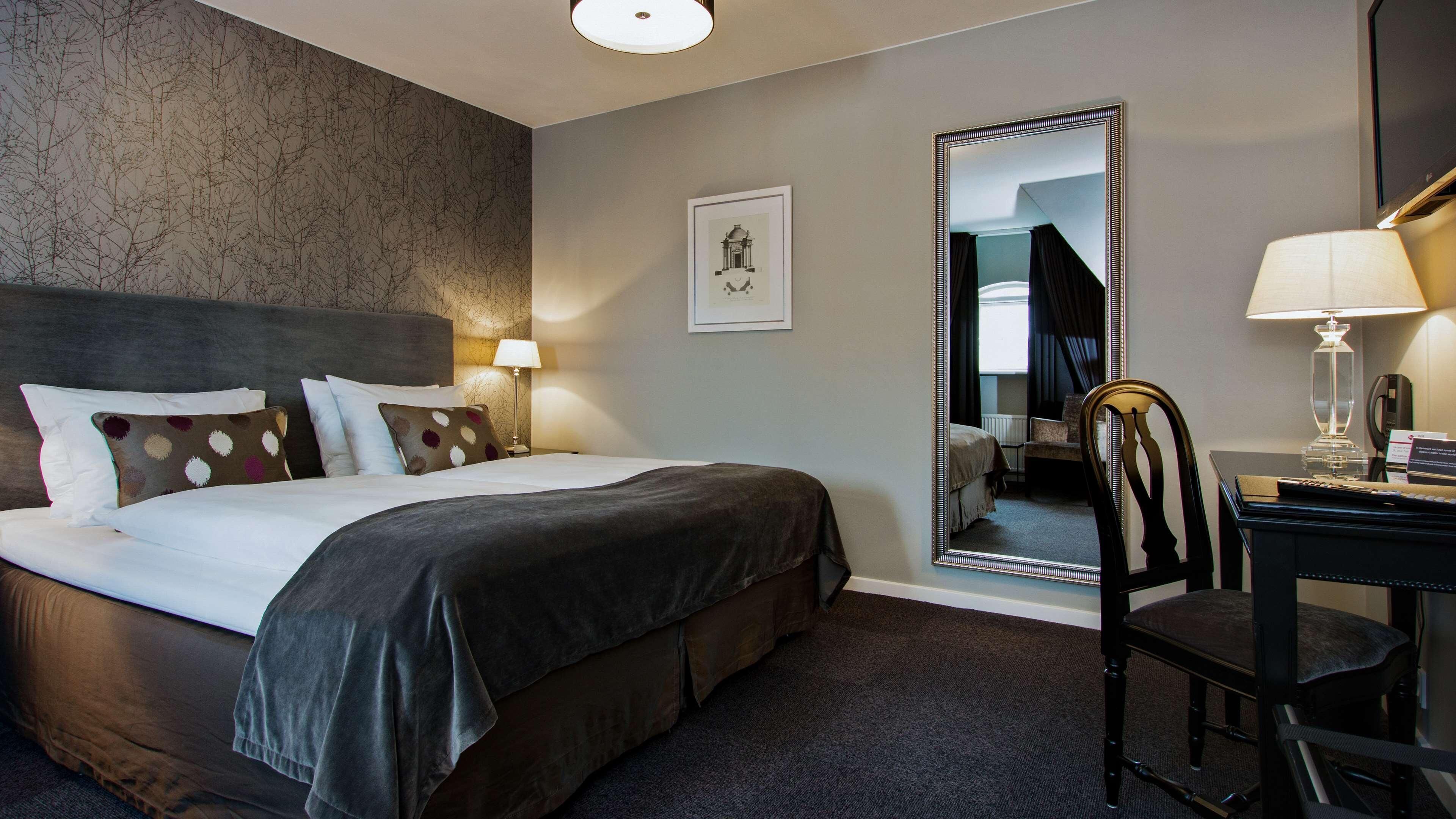 A hotel room with a bed, a desk, a chair, a mirror, and a picture on the wall.