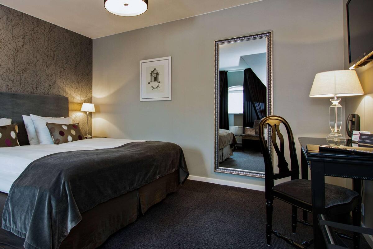 A hotel room with a large bed, a desk, a chair, a mirror, and a lamp.
