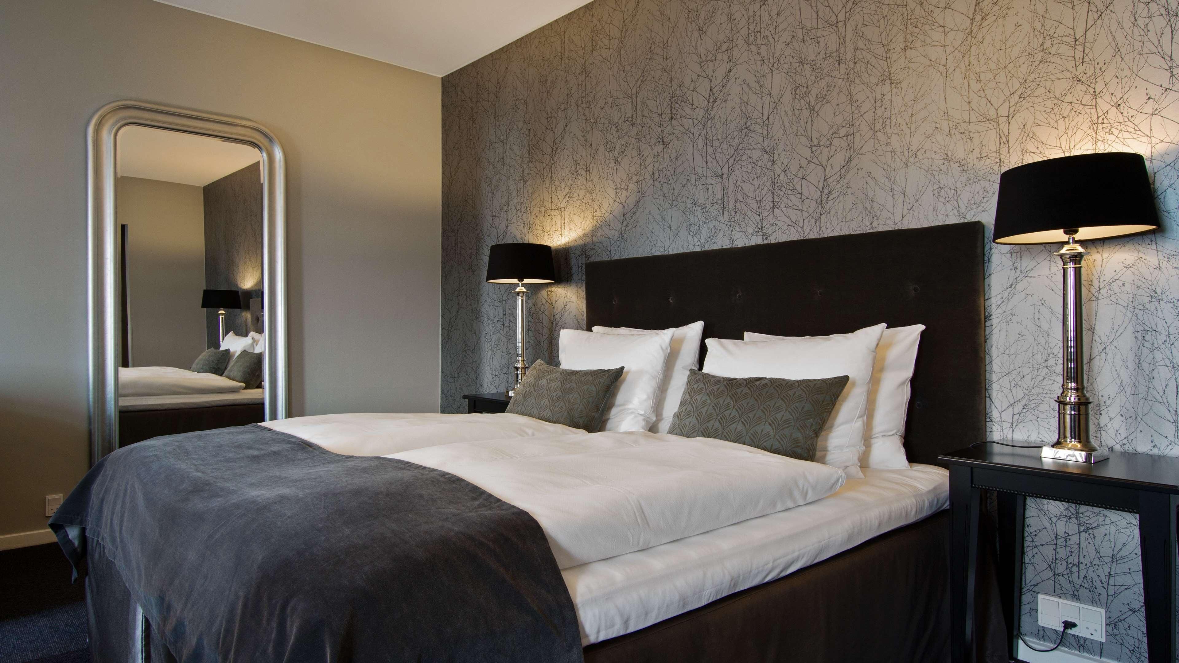A neatly made bed with white and dark bedding, two bedside lamps, a large mirror, and a wall with a tree branch pattern.
