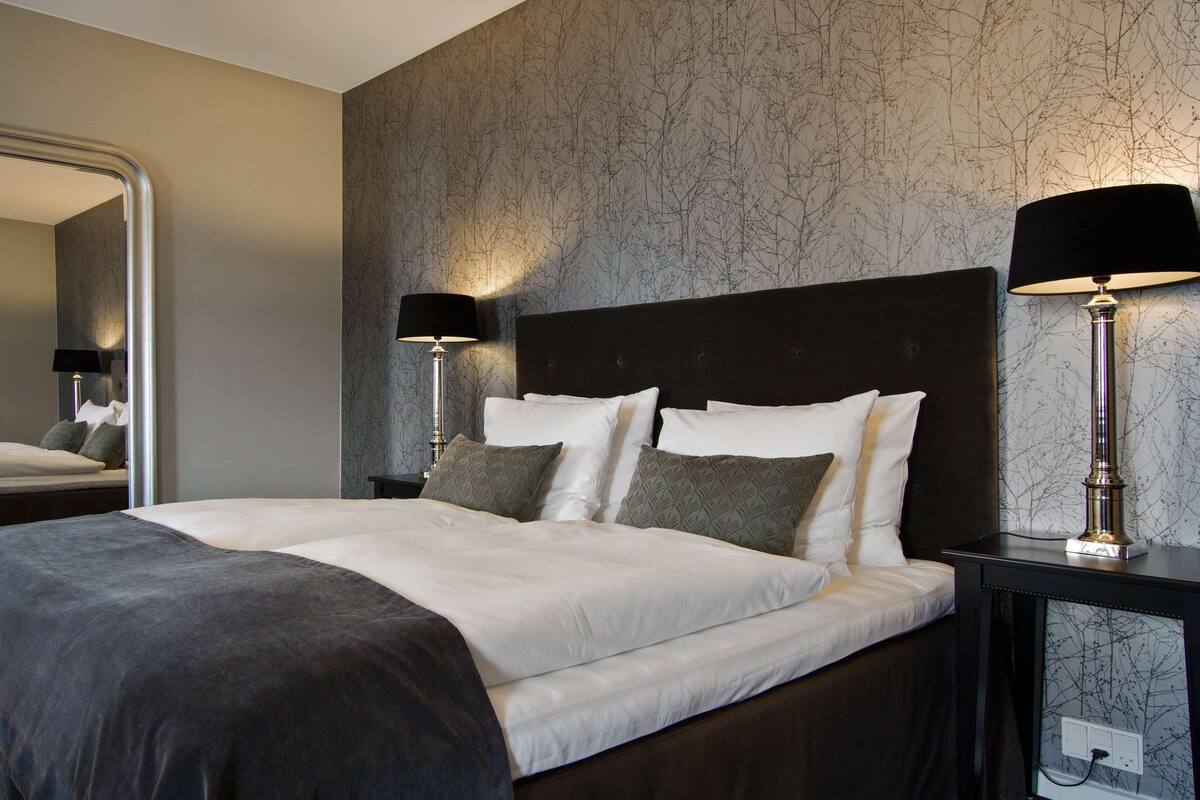 A neatly made bed with white and gray bedding, two bedside lamps, and a large mirror on the left.