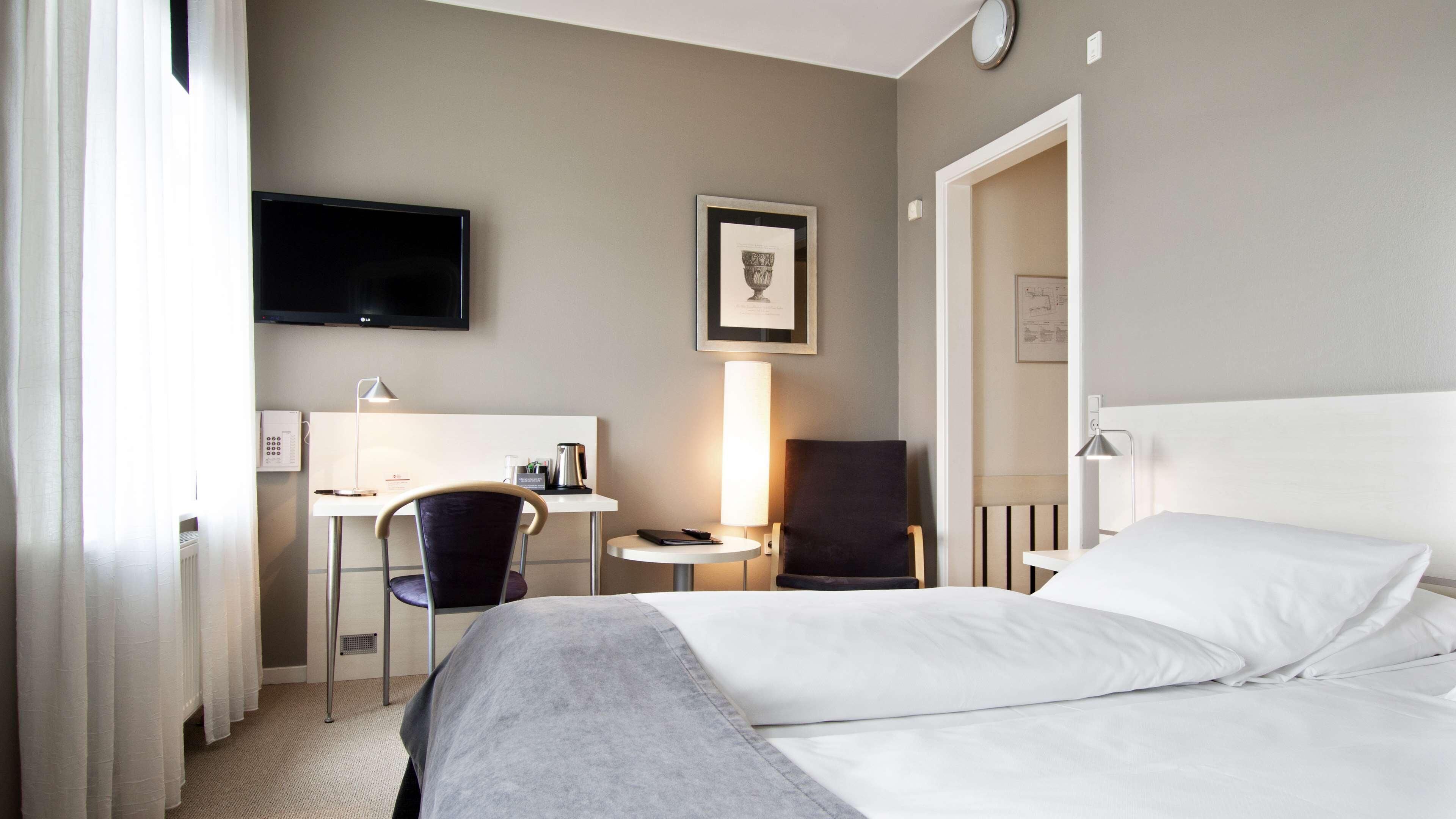 A hotel room with a bed, a desk with a chair, a TV, and a lamp.