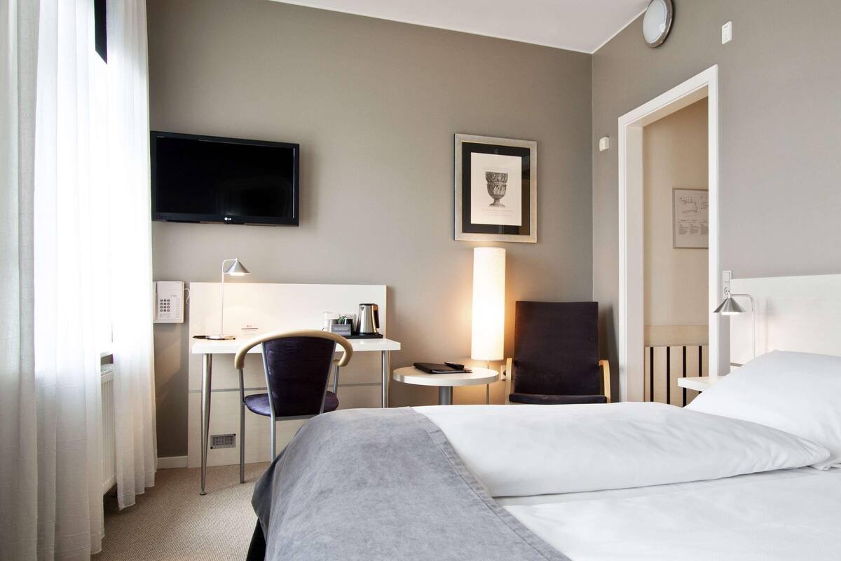 A hotel room with a bed, a desk with a chair, a TV, and a lamp.