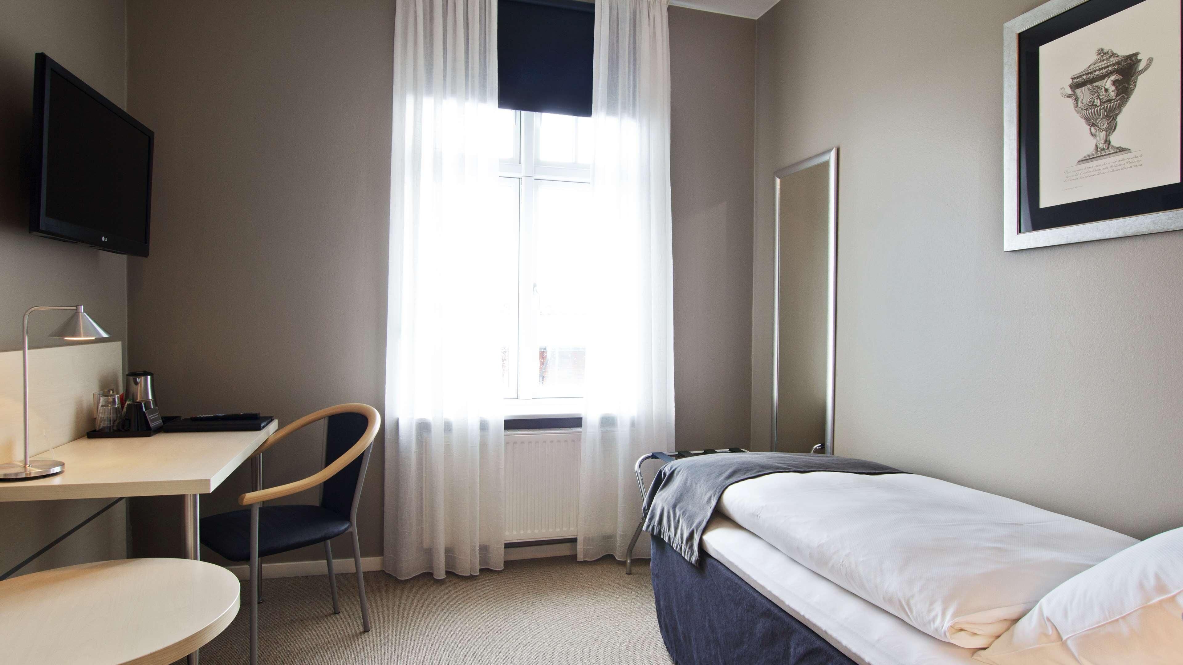A hotel room with a bed, a desk, a chair, a television, a window with curtains, and a framed picture on the wall.