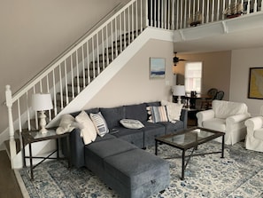 A living room with a gray sectional sofa, a glass coffee table, a white armchair, and a staircase with white railings.