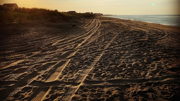 Una spiaggia con solchi di pneumatici che conducono all'acqua, cielo sereno e edifici sullo sfondo.