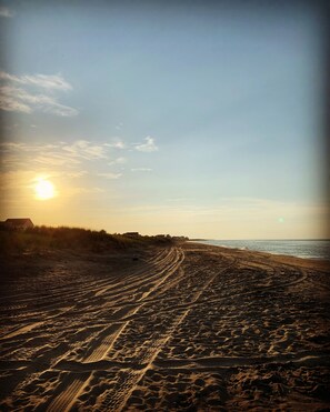 A beach with tire tracks leading to the water, a clear sky, and buildings in the background.