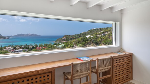 A room with a large window overlooking a coastal landscape, a wooden desk, and two wicker chairs.