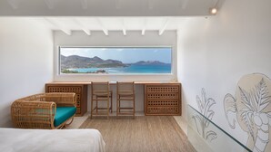 A bedroom with a large window overlooking a scenic coastal view, a wooden desk with two chairs, and a bed with a white headboard.