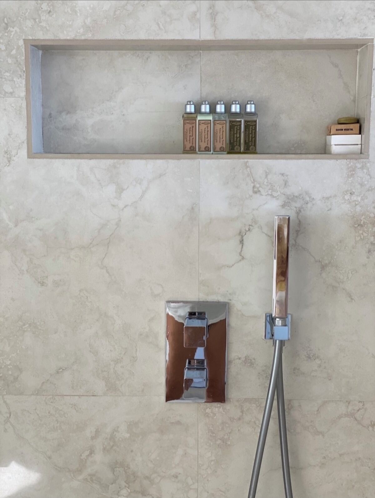 Un baño con suelo de mármol, cabezal de ducha cromado y un estante con artículos de aseo.