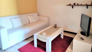 Ein Wohnzimmer mit einem weißen Sofa, einem kleinen Couchtisch, einem roten Teppich, einem Fernseher und einem Regal mit dekorativen Gegenständen.