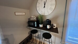 A kitchen with a large wall clock, a microwave, a coffee maker, two stools, and a potted plant.