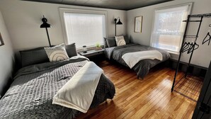 A room with two beds, wooden flooring, and large windows with blinds.