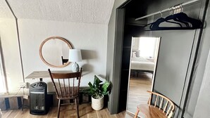 A compact room with a wooden floor, a round mirror, a small table, two chairs, a potted plant, and a heater.