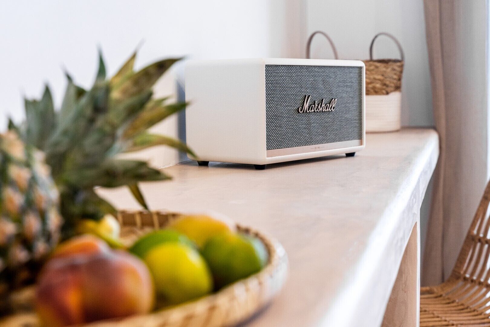 A white Marshall speaker on a light-colored countertop with a wicker basket of fruits and a wicker chair in the background.