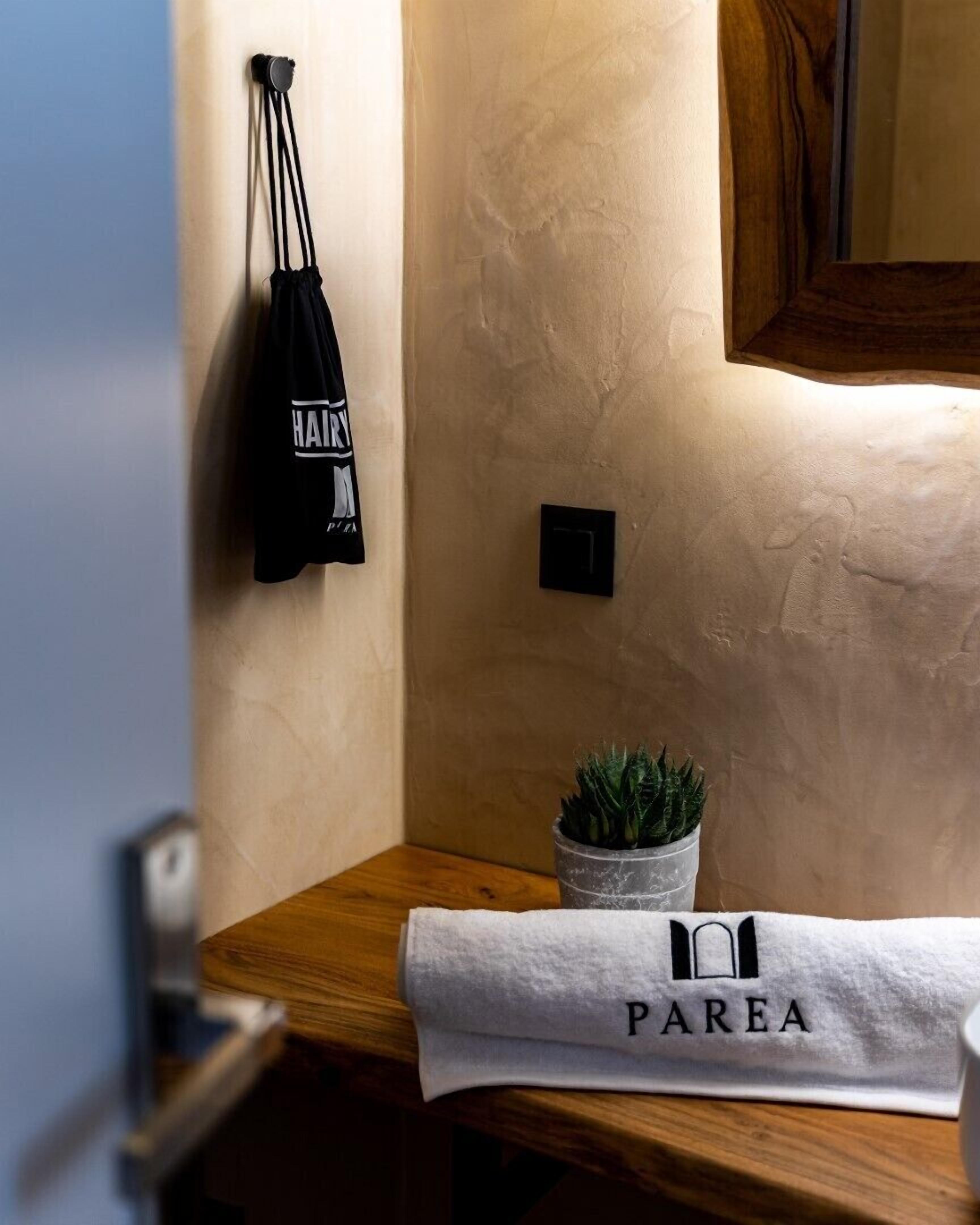 Une salle de bain avec un plan de toilette en bois, une serviette ornée du mot « PAREA », une petite plante en pot et un sac noir accroché au mur.
