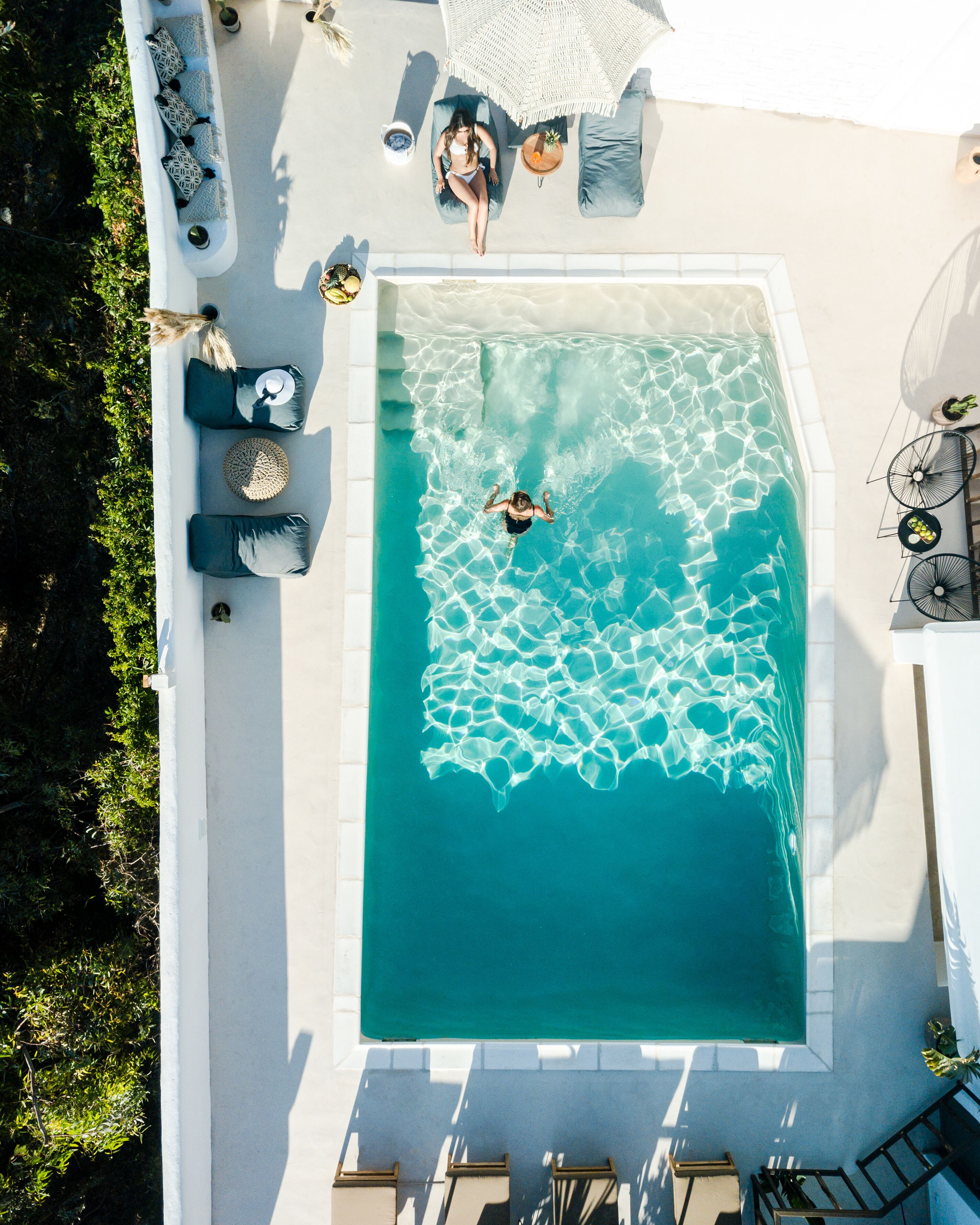 An aerial view of a rectangular swimming pool with clear blue water, surrounded by a white deck with lounge chairs and a small table with a drink.