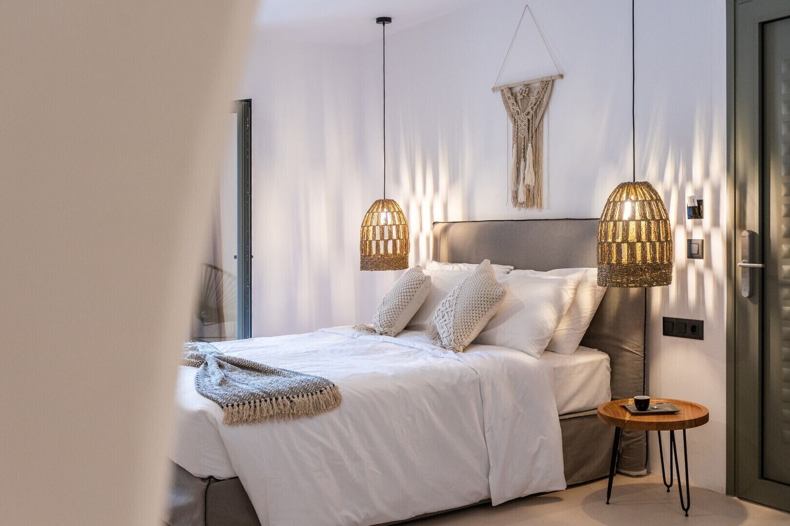 Un dormitorio moderno con una cama grande, dos lámparas colgantes, un colgador de macramé en la pared y una mesita de madera pequeña.
