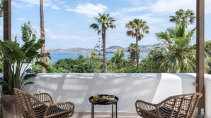 Un balcon avec des fauteuils en osier, une petite table sur laquelle est disposé un plateau de fruits, et une vue sur la mer et les palmiers.