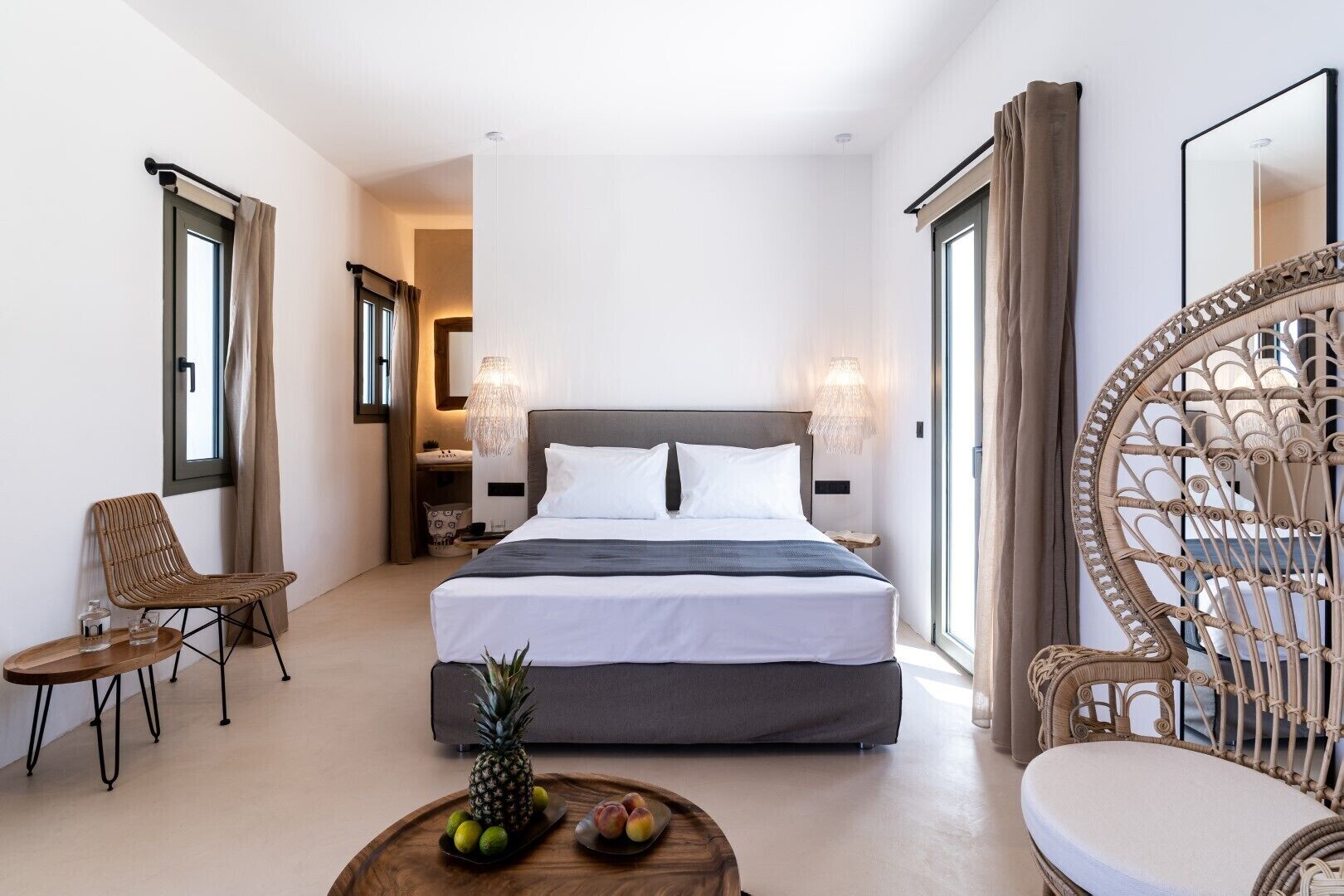 A modern hotel room with a large bed, a small table with fruits, a wicker chair, and a large mirror.