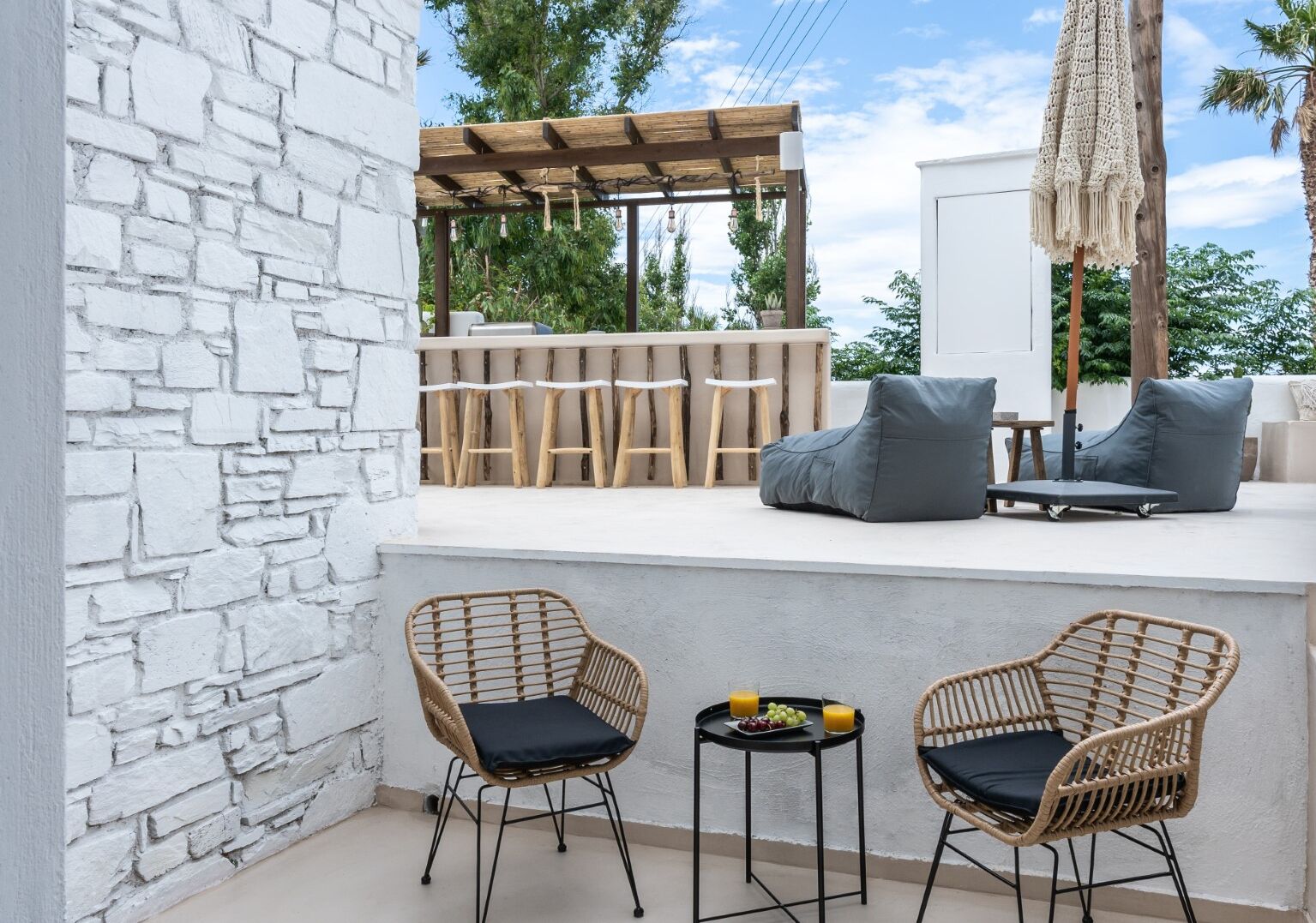 Un patio con sillas de mimbre, una mesa pequeña con frutas y bebidas, y una zona de bar con taburetes.