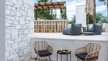 Un patio con sillas de mimbre, una mesa pequeña con frutas y bebidas, y una zona de bar con taburetes.