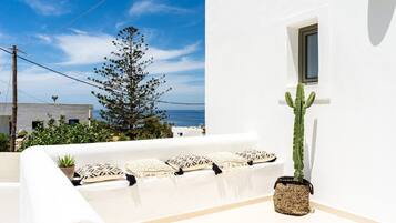 A white outdoor seating area with cushions, a cactus plant, and a view of the sea and buildings in the background.