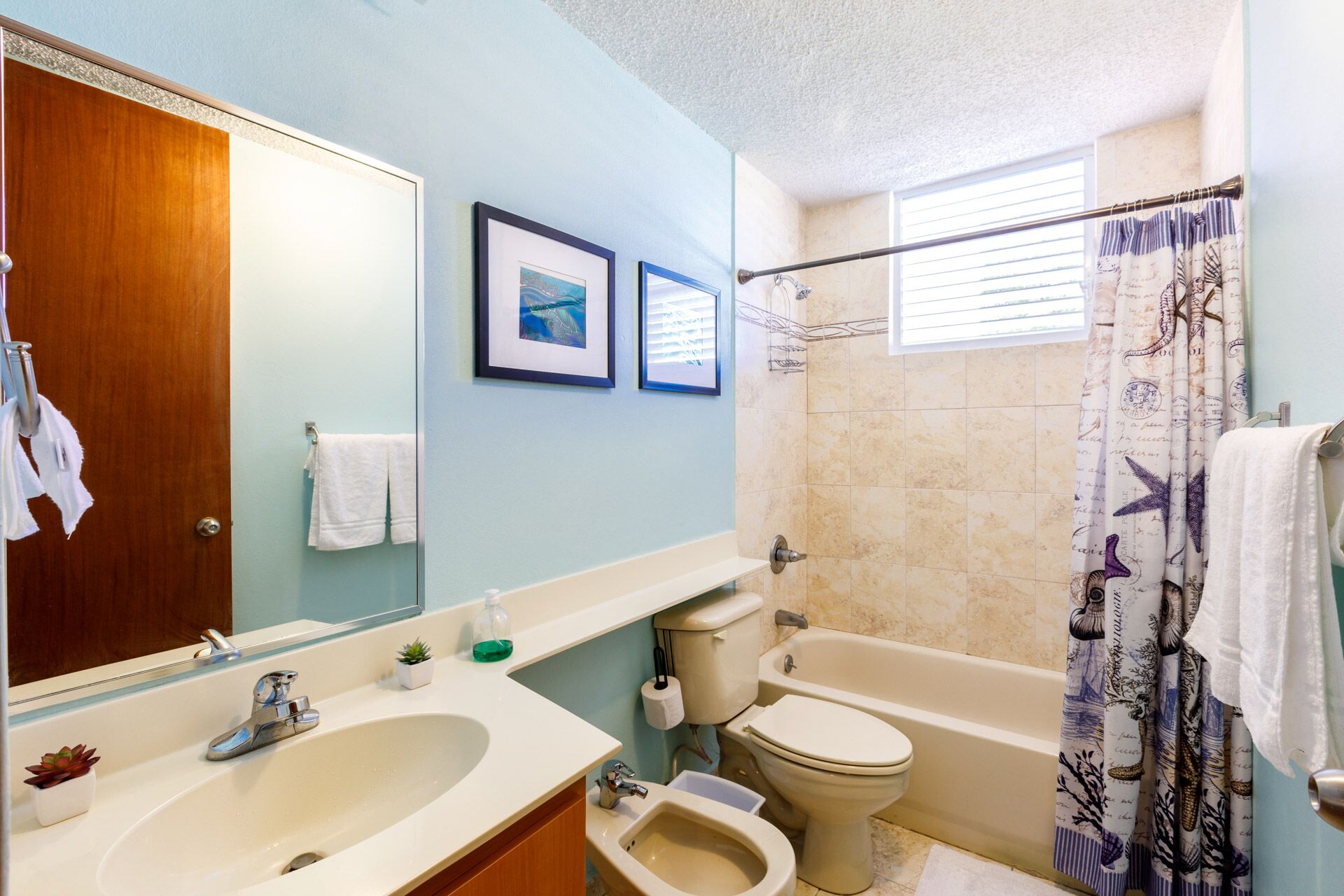 A bathroom with a sink, mirror, toilet, and bathtub. There are framed pictures on the wall and a shower curtain with a nautical theme.