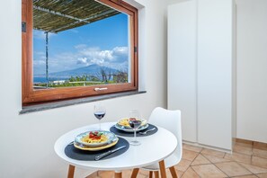 Ein Essbereich mit einem Tisch für zwei Personen, zwei Pasta-Tellern, zwei Gläsern Rotwein und Blick auf eine Berglandschaft durch ein Fenster.