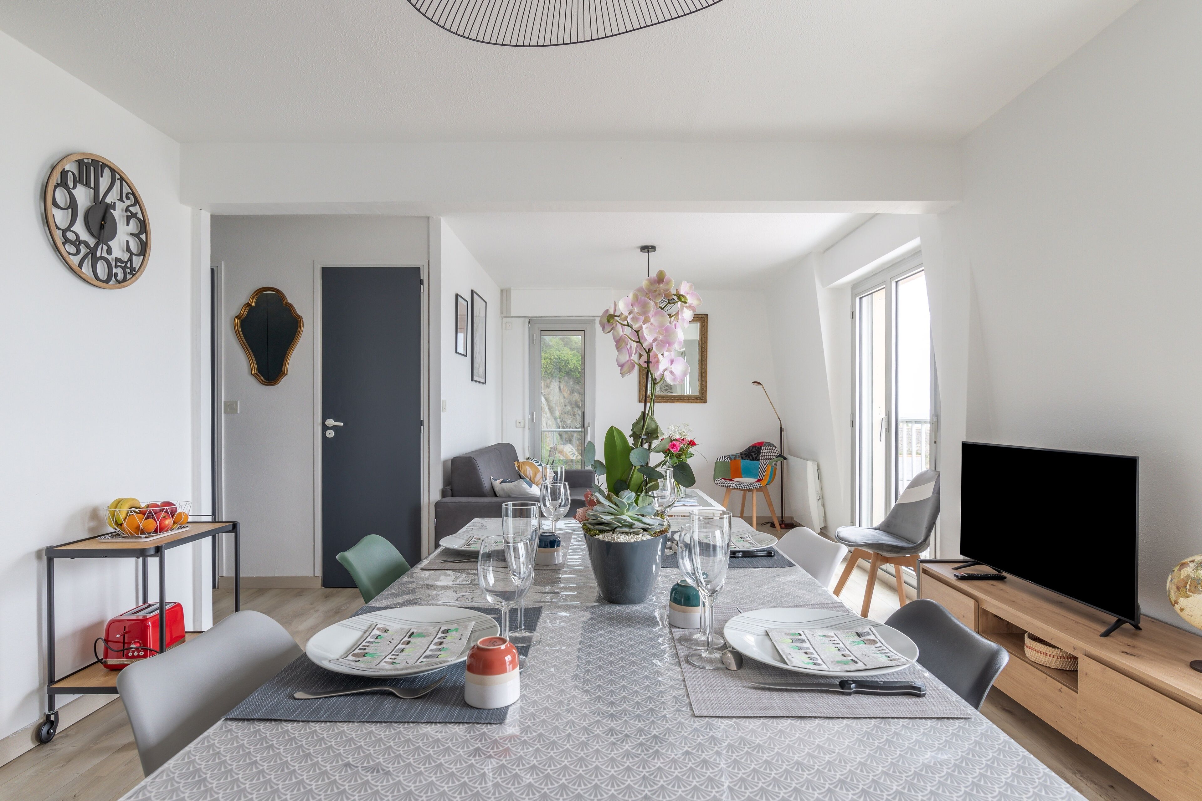 Un espace repas moderne avec une longue table dressée pour six personnes, un arrangement floral en pot au centre et un téléviseur posé sur un meuble en bois.