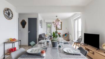 Un espace repas moderne avec une longue table dressée pour six personnes, un arrangement floral en pot au centre et un téléviseur posé sur un meuble en bois.