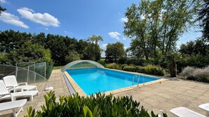 A swimming pool with clear blue water, surrounded by greenery and trees. There are lounge chairs and a covered seating area. - Surfinia - piscine partagée - proche Europa Park (Hessenheim)