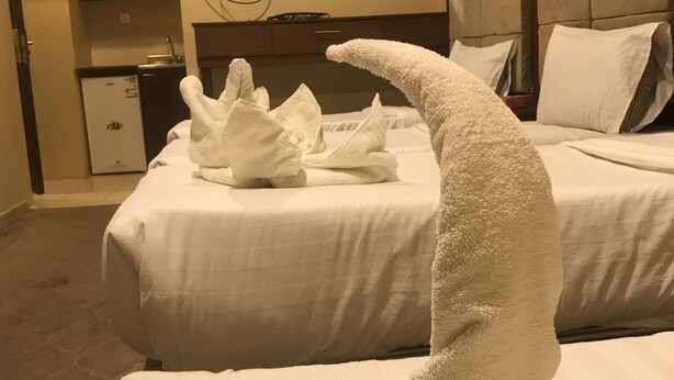 Een hotelkamer met twee bedden, een aan de muur bevestigde televisie en een kleine kitchenette op de achtergrond.